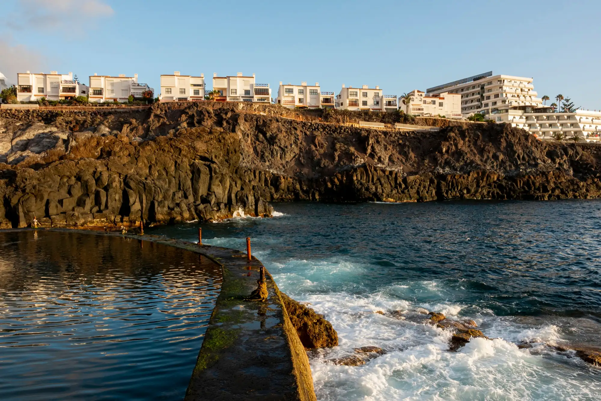 Costa rochosa de origem vulcânica com moradias e um hotel brancos no topo e, em primeiro plano, o mar com ondas e uma piscina natural